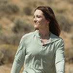 A woman wearing a sage green long-sleeve henley shirt walking outdoors in a dry, brush-filled landscape.