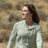 A woman wearing a sage green long-sleeve henley shirt walking outdoors in a dry, brush-filled landscape.