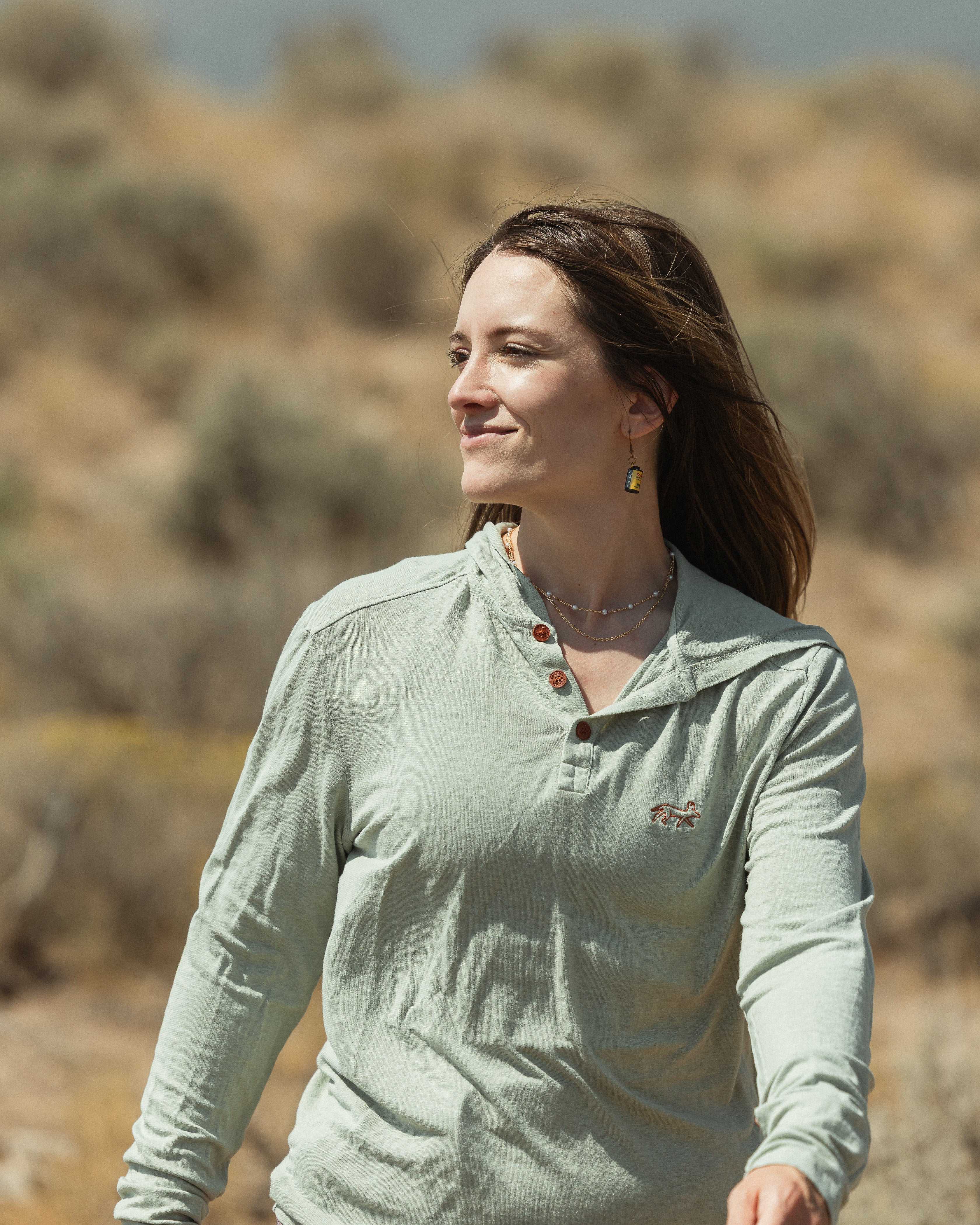 A woman wearing a sage green long-sleeve henley shirt walking outdoors in a dry, brush-filled landscape.