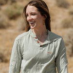 A woman smiling outdoors in a light sage green long-sleeve henley shirt with brown buttons and a small embroidered logo on the left chest, set against a blurred natural background.