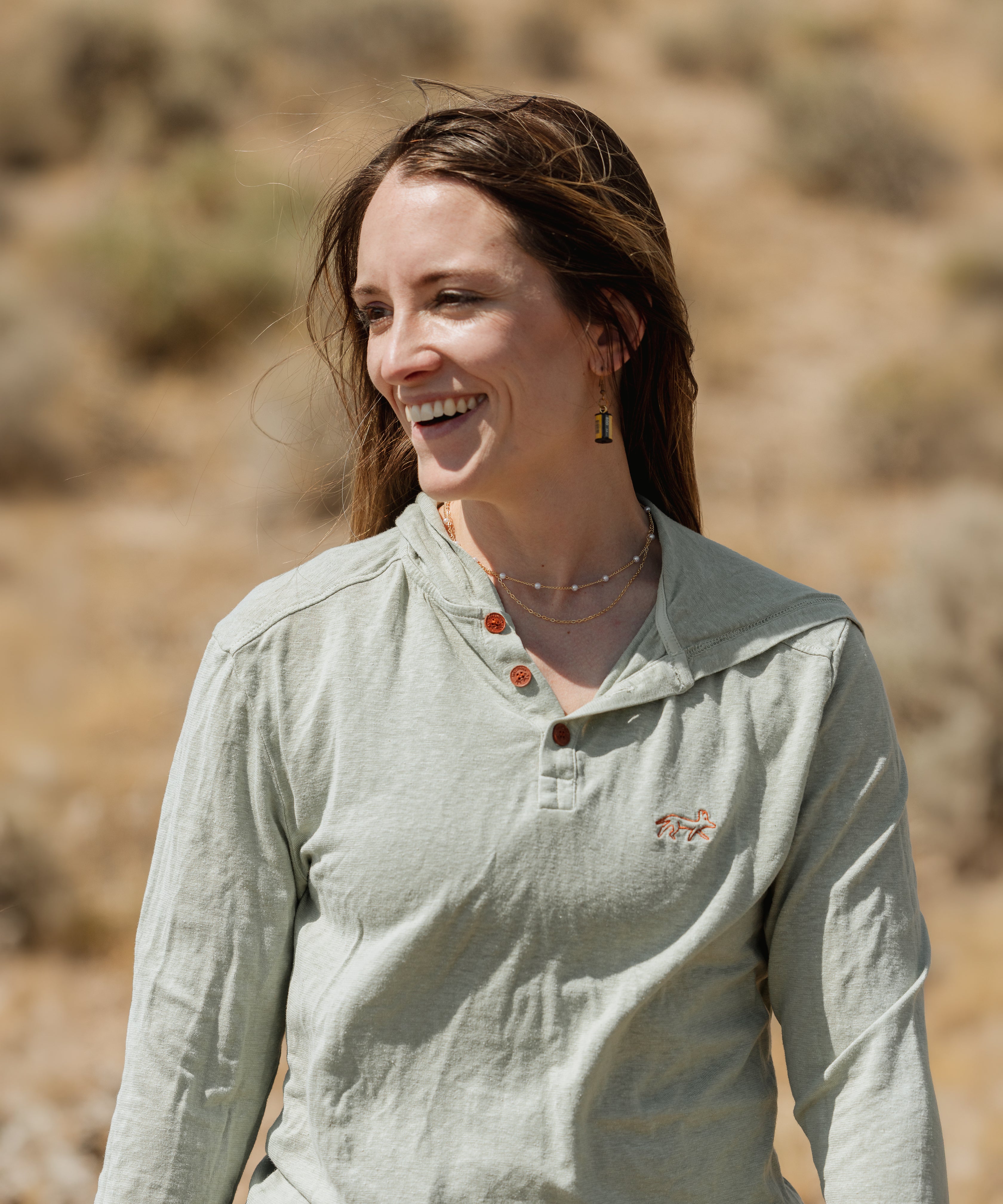 A woman smiling outdoors in a light sage green long-sleeve henley shirt with brown buttons and a small embroidered logo on the left chest, set against a blurred natural background.