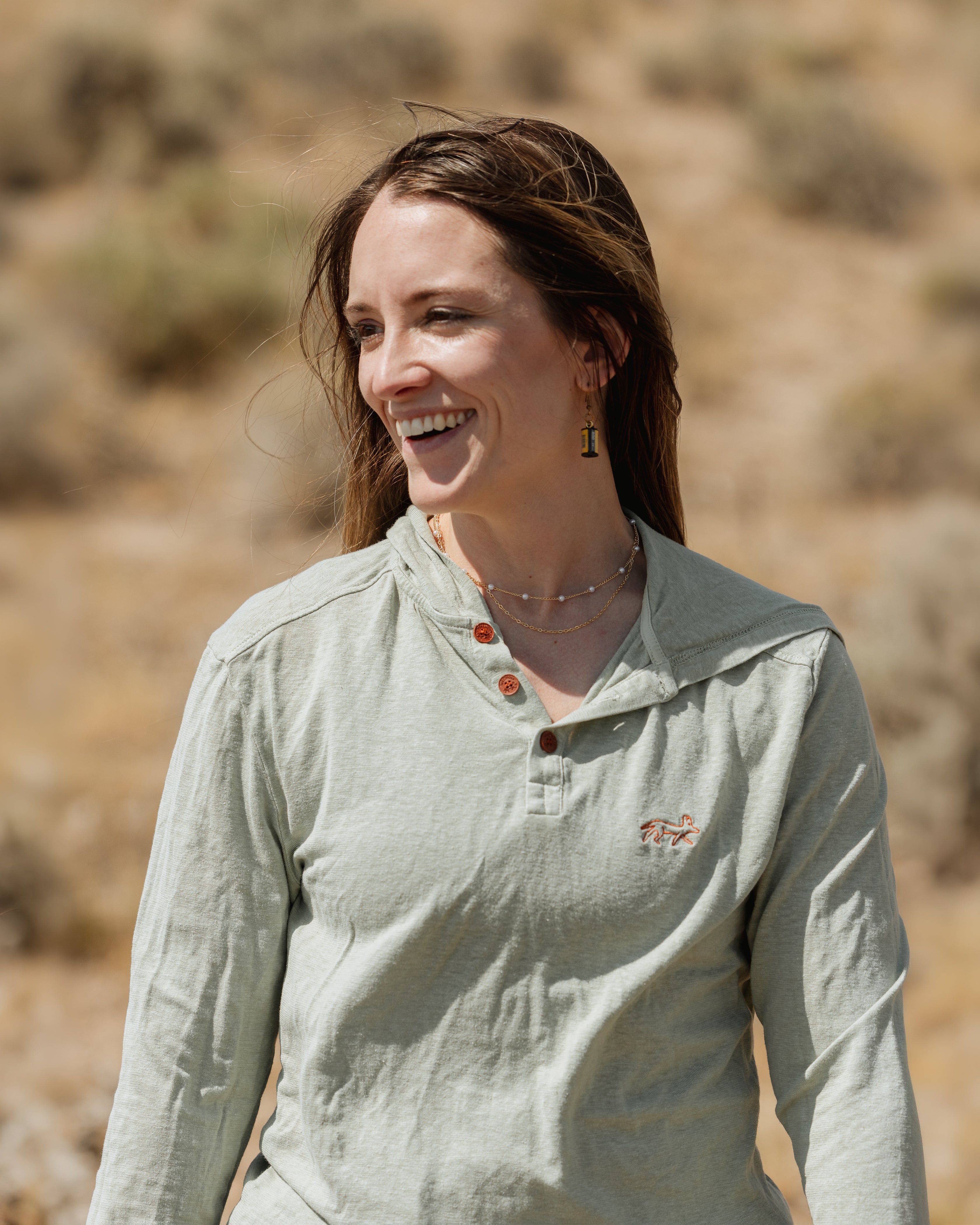 A woman smiling outdoors in a light sage green long-sleeve henley shirt with brown buttons and a small embroidered logo on the left chest, set against a blurred natural background.