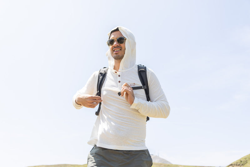 Man wearing a white hoodie with a hood, sunglasses, and carrying a backpack against a clear blue sky.