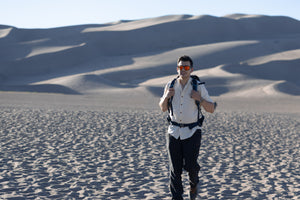 Man with a backpack in the dunes with a scenic view wearing Everglades - Men's Short-Sleeve Shirt in Chamisa (Tan) (front view)