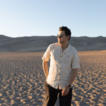 Man with a backpack in the dunes with a scenic view wearing Everglades - Men's Short-Sleeve Shirt in Chamisa (Tan) (front view)