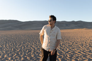 Man with a backpack in the dunes with a scenic view wearing Everglades - Men's Short-Sleeve Shirt in Chamisa (Tan) (front view)