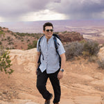 Man with a backpack standing on a rocky landscape with a scenic view wearing Everglades - Men's Short-Sleeve Shirt in Blue Sage (Light Blue) (front view)