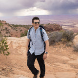 Man with a backpack standing on a rocky landscape with a scenic view wearing Everglades - Men's Short-Sleeve Shirt in Blue Sage (Light Blue) (front view)
