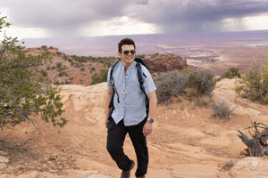 Man with a backpack standing on a rocky landscape with a scenic view wearing Everglades - Men's Short-Sleeve Shirt in Blue Sage (Light Blue) (front view)