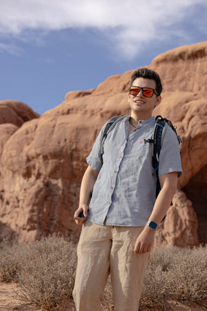 Man with a backpack standing on a rocky landscape with a scenic view wearing Everglades - Men's Short-Sleeve Shirt in Blue Sage (Light Blue) (front view)