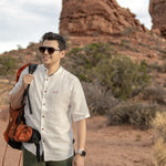 Man with a backpack standing on a rocky landscape with a scenic view wearing Everglades - Men's Short-Sleeve Shirt in Chamisa (Tan) (front view)