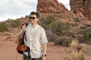 Man with a backpack standing on a rocky landscape with a scenic view wearing Everglades - Men's Short-Sleeve Shirt in Chamisa (Tan) (front view)