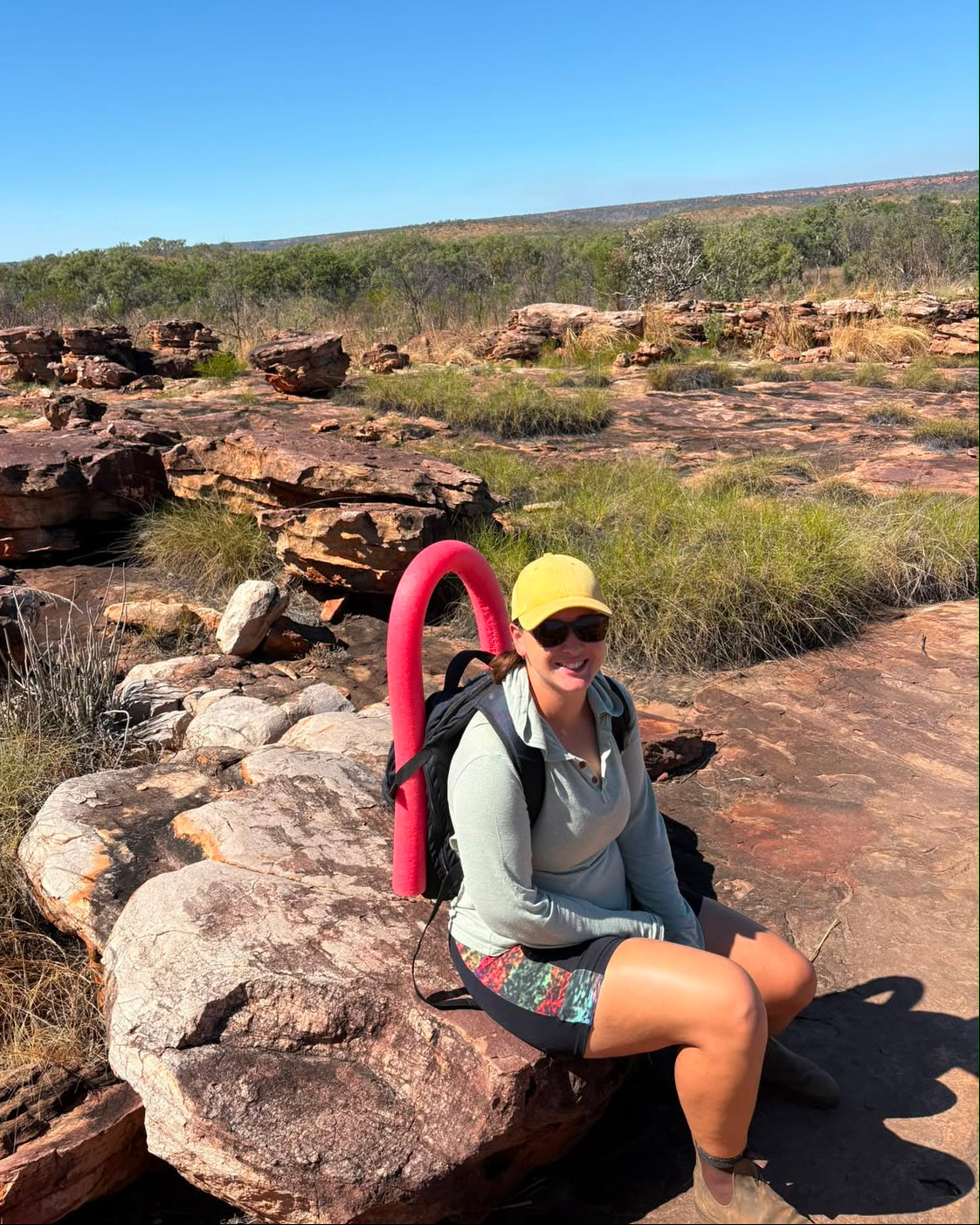 A woman sitting on a large rock in a rocky, scrub-covered outdoor landscape under a clear blue sky, wearing a yellow cap, sunglasses, a light gray long-sleeve top, and shorts, with a bright pink curved object behind her.