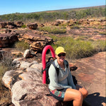 A woman sitting on a large rock in a rocky, scrub-covered outdoor landscape under a clear blue sky, wearing a yellow cap, sunglasses, a light gray long-sleeve top, and shorts, with a bright pink curved object behind her.