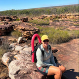 A woman sitting on a large rock in a rocky, scrub-covered outdoor landscape under a clear blue sky, wearing a yellow cap, sunglasses, a light gray long-sleeve top, and shorts, with a bright pink curved object behind her.